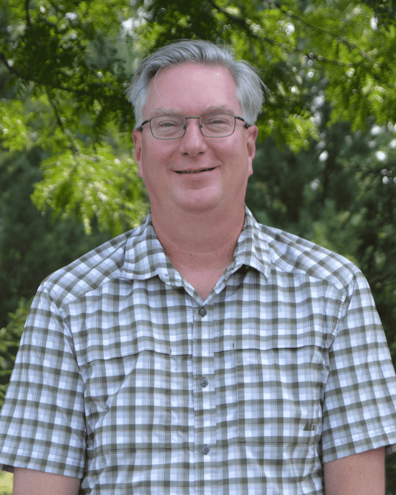 Portrait of a smiling man with gray hair and glasses wearing a plaid short-sleeve shirt, standing outdoors with trees in the background.